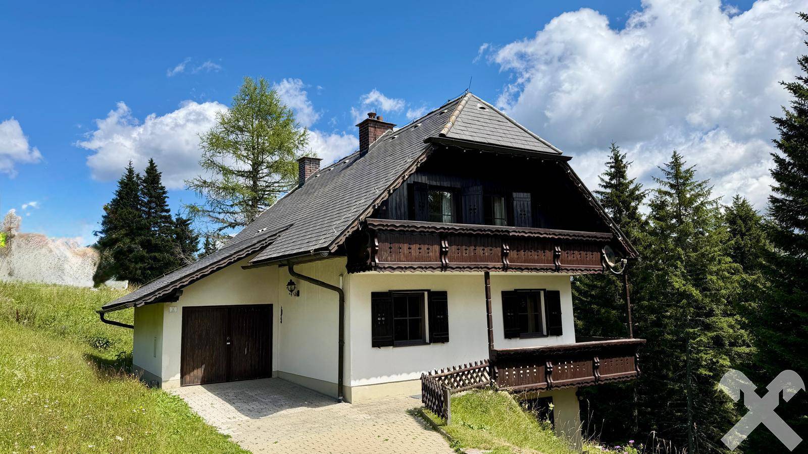 Alpines Haus mit Satteldach und integrierter Garage, umgeben von üppiger Natur.