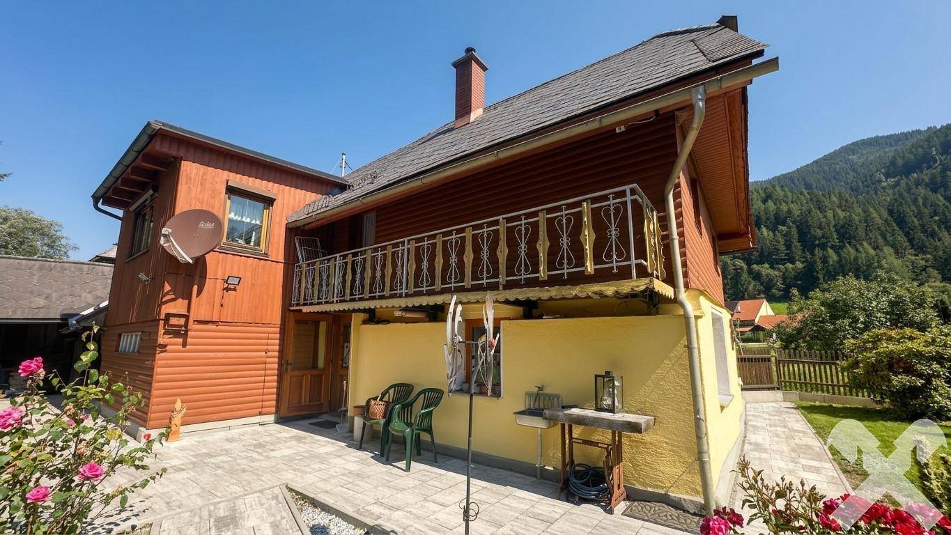 Seitenansicht des Hauses mit traditionellem Balkon und gepflasterter Terrasse, umgeben von grüner Natur.