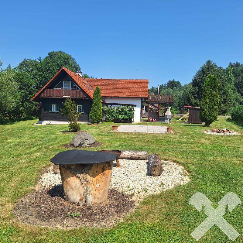 Weitläufiger Garten mit Einfamilienhaus, Feuerstelle und viel Grünfläche an einem sonnigen Tag.