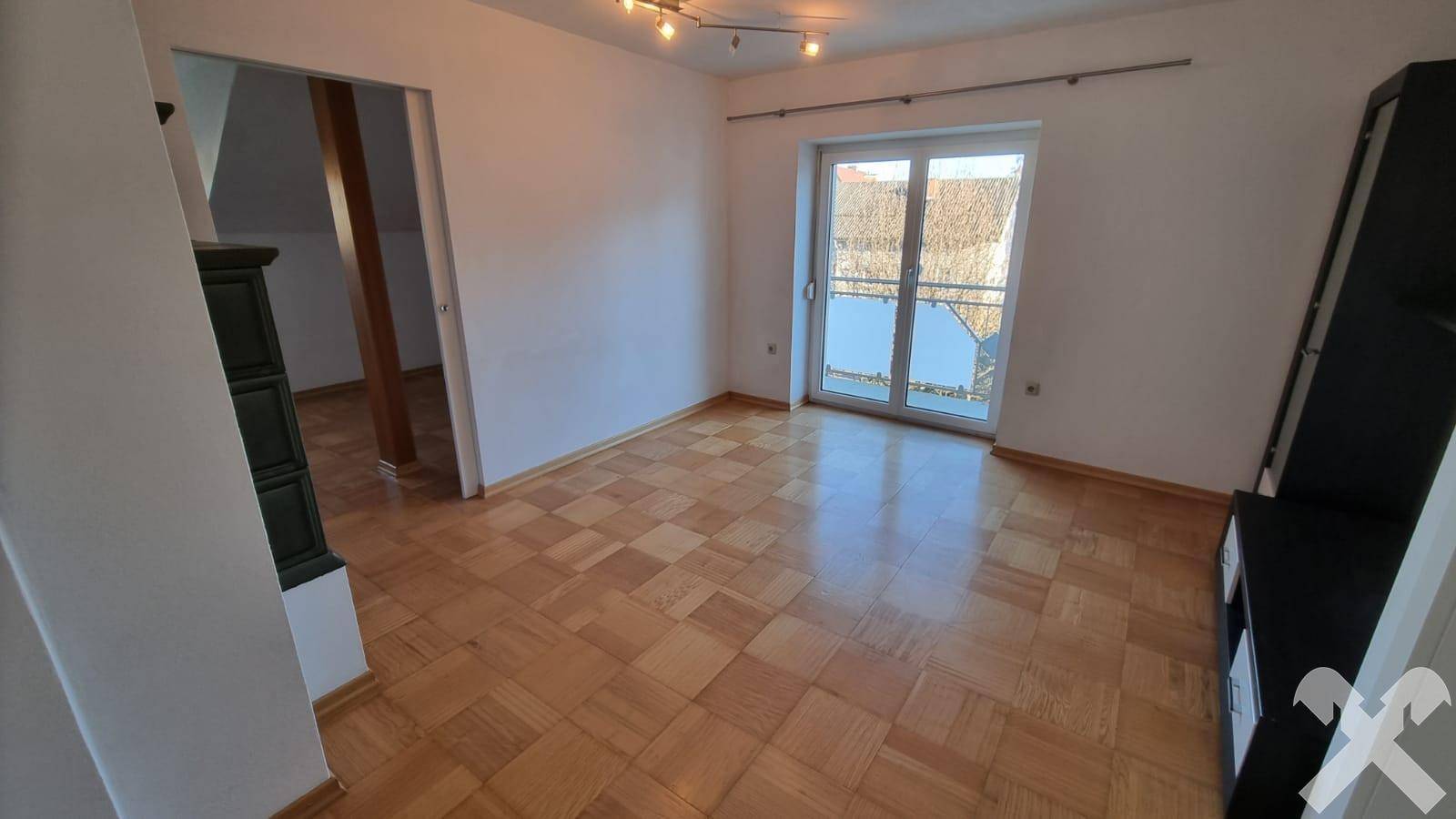 Heller Wohnraum mit Parkettboden, Zugang zum Balkon und einem traditionellen Kachelofen.
