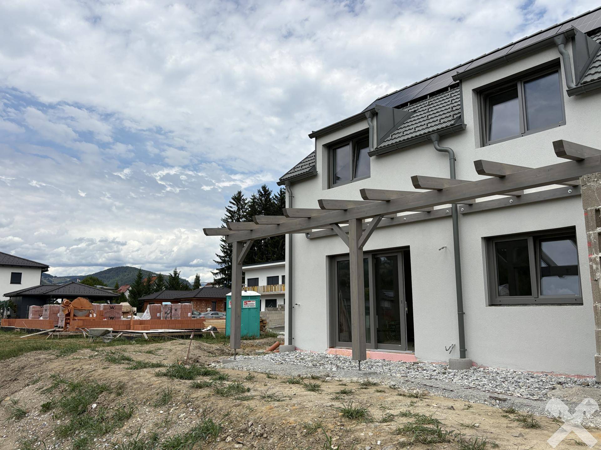 Neubau-Doppelhaushälfte mit Pergola-Struktur und Blick auf die umliegende Baustelle und Hügellandschaft.