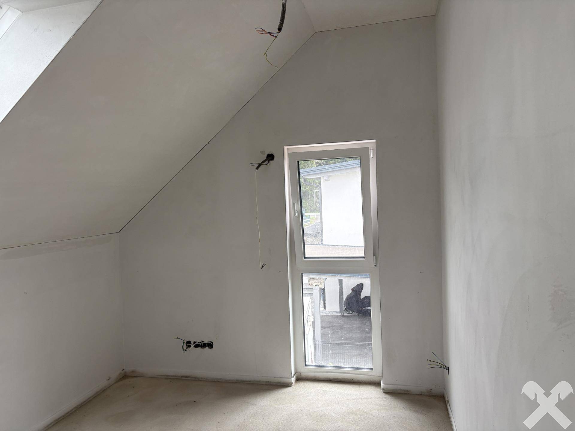Unfertiges Dachzimmer mit schräger Decke und hohem Fenster, das viel natürliches Licht hereinlässt.