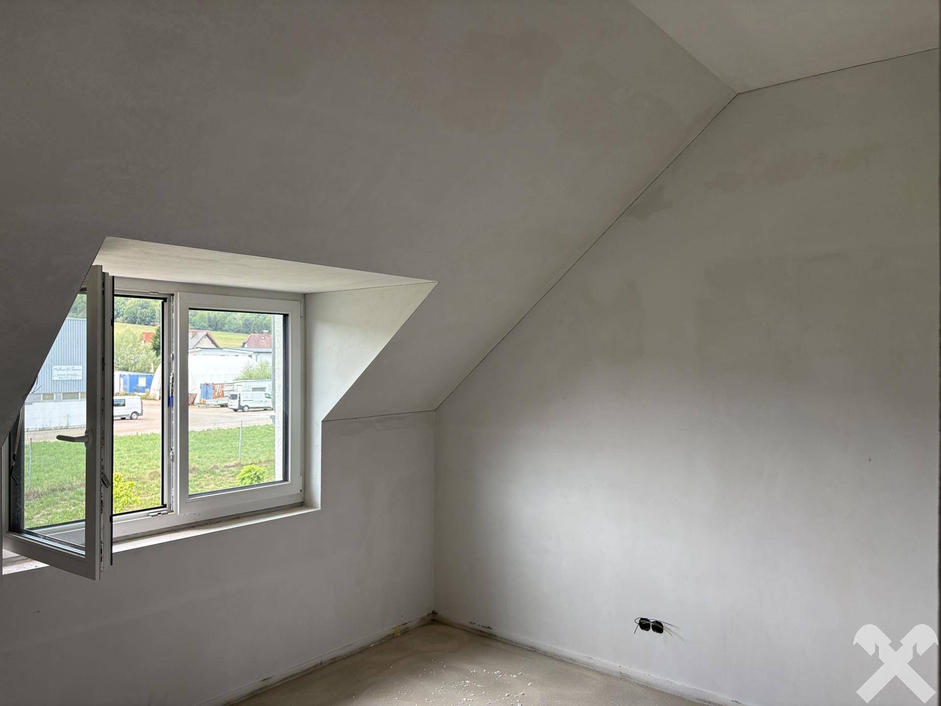 Unfertiges Dachzimmer mit schräger Decke und Fensterblick ins Grüne und auf umliegende Gebäude.