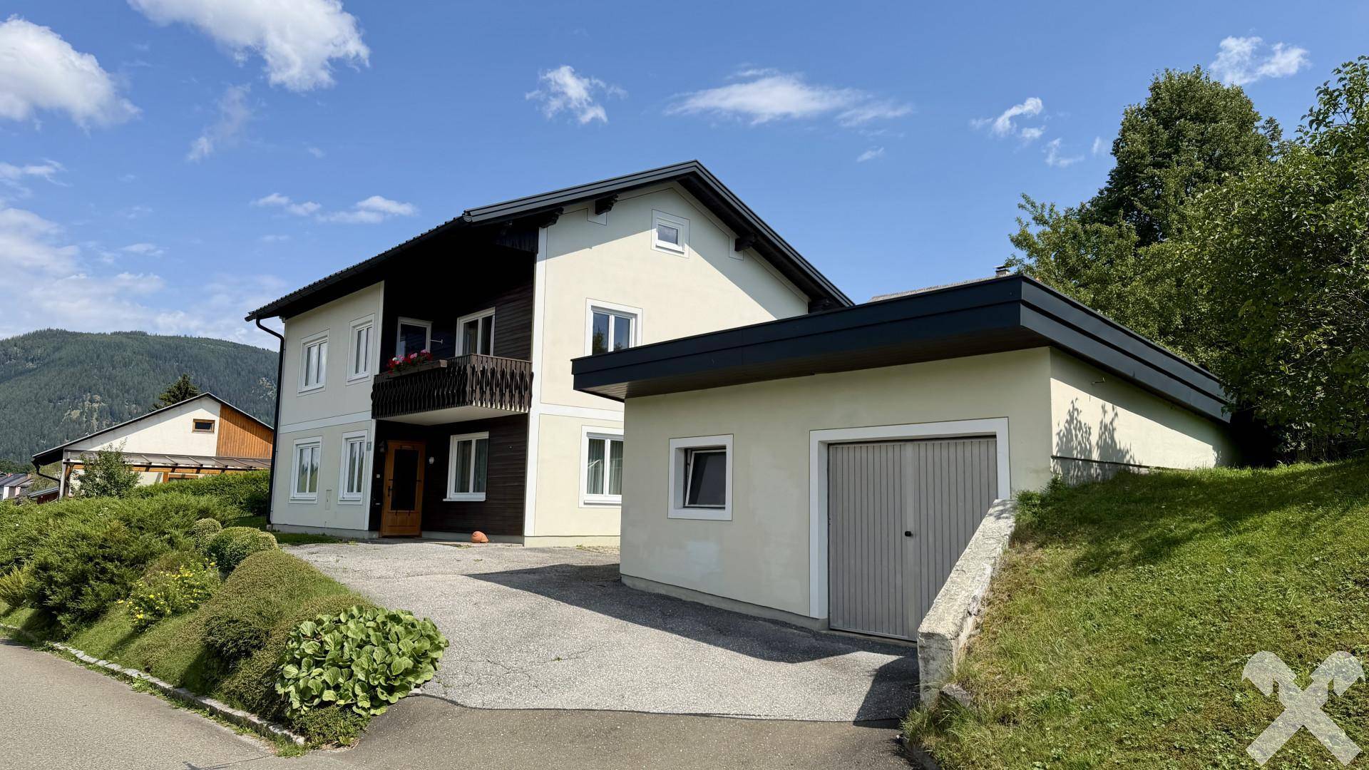 Ansicht des Hauses mit Garage und Zufahrt, eingebettet in eine grüne Hügellandschaft.