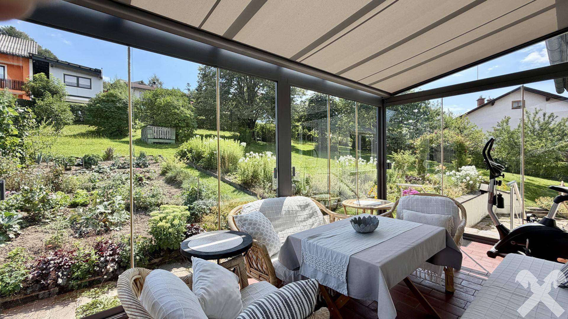 Heller Wintergarten mit Glaswänden, Rattanmöbeln und Blick auf den gepflegten Garten.