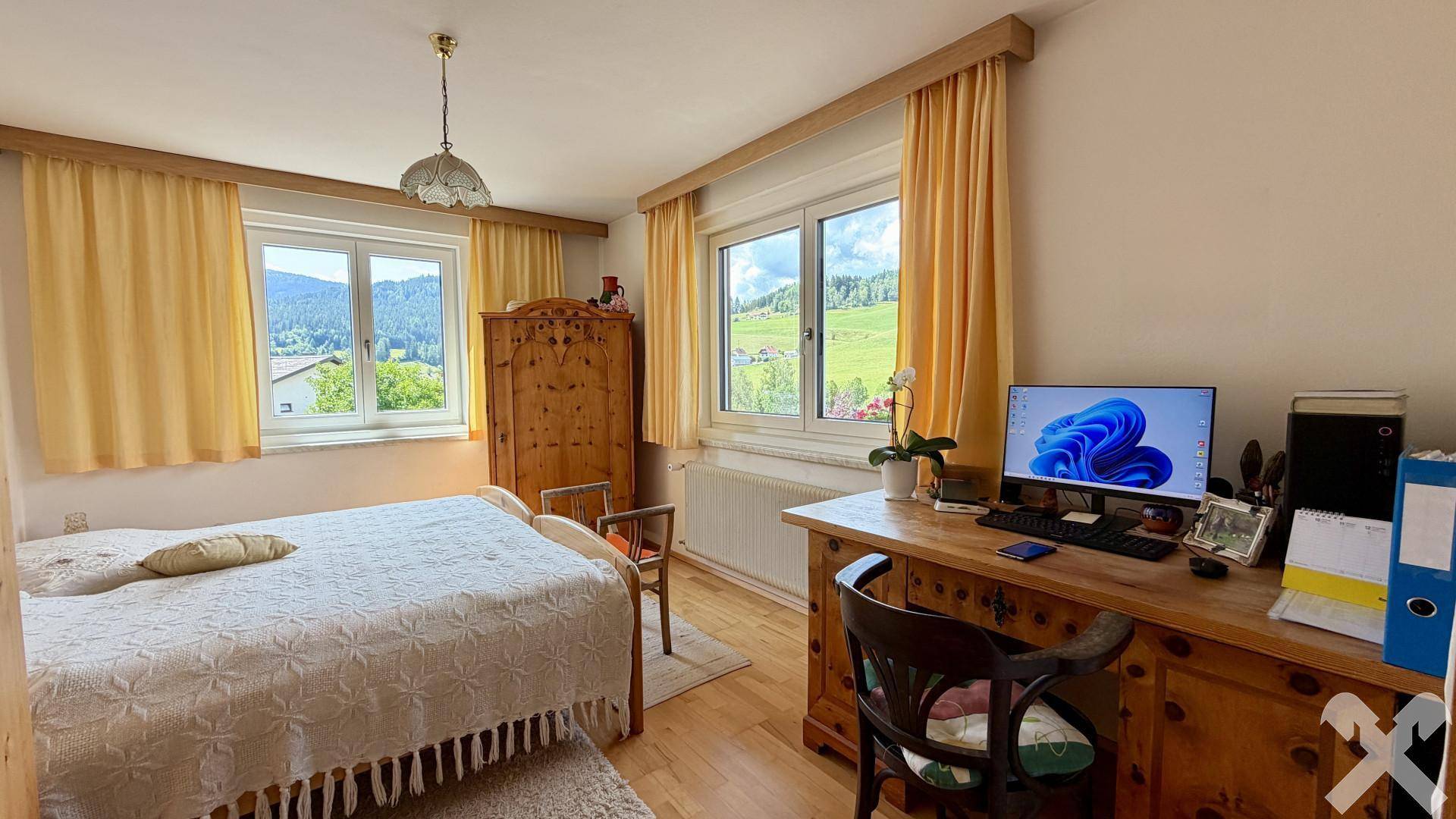 Helles Schlafzimmer mit Holzmöbeln, Schreibtisch und Fenstern mit Bergblick.