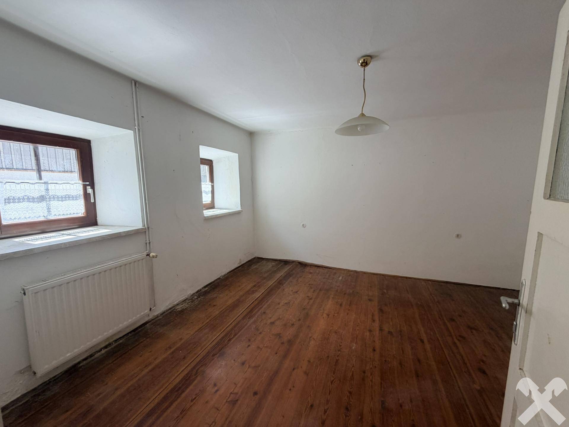 Leeres Zimmer mit Holzdielenboden, weißen Wänden und einem Fenster mit Heizkörper.