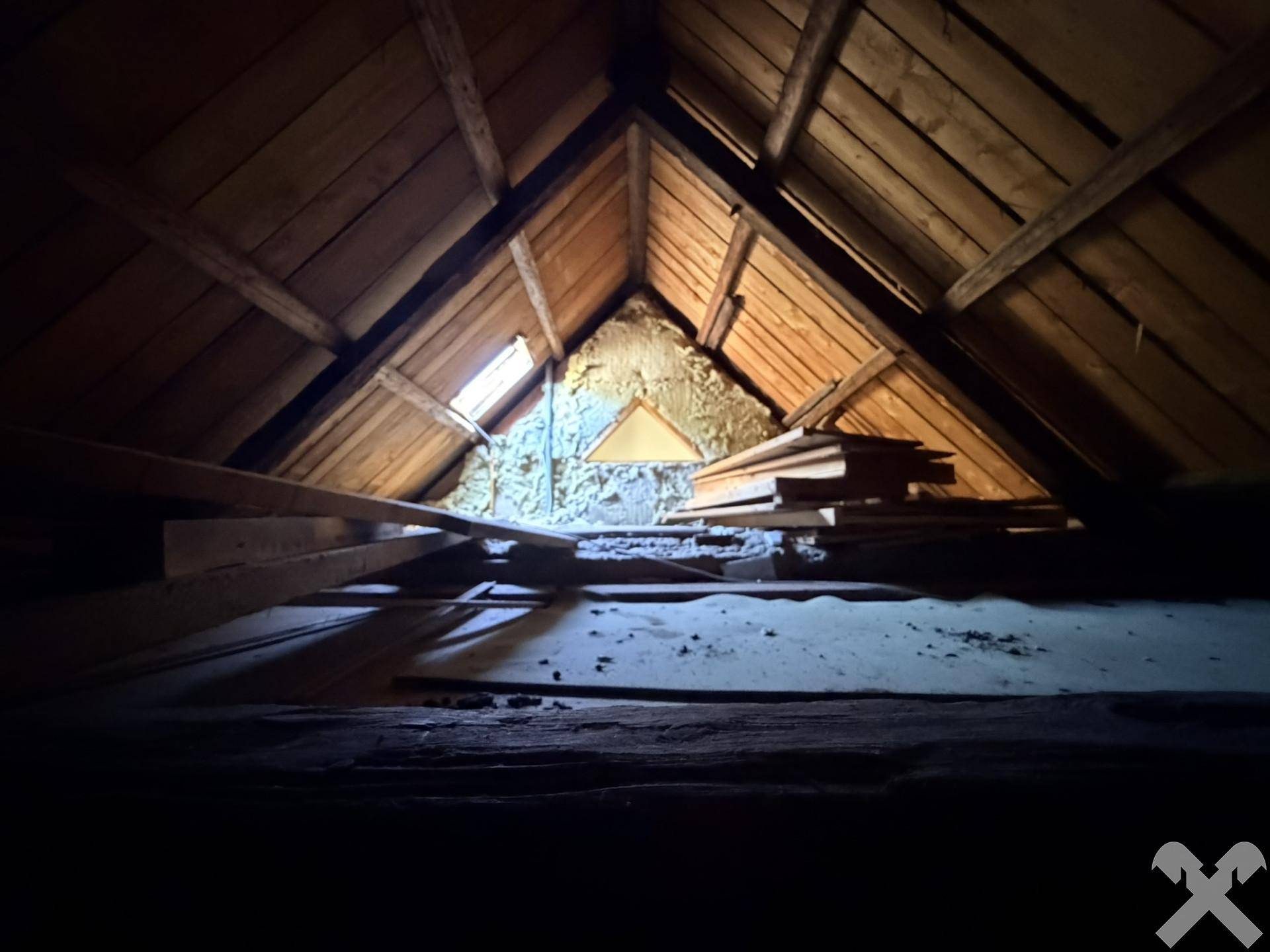Dunkler, unfertiger Dachboden mit sichtbaren Holzbalken und einem kleinen Dachfenster, bereit für den Ausbau.