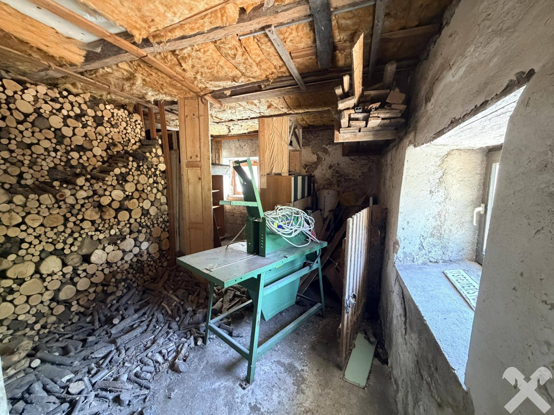 Rustikaler Abstellraum mit großem Holzstapel, einer alten Maschine und einem kleinen Fenster, ideal für Lagerung.