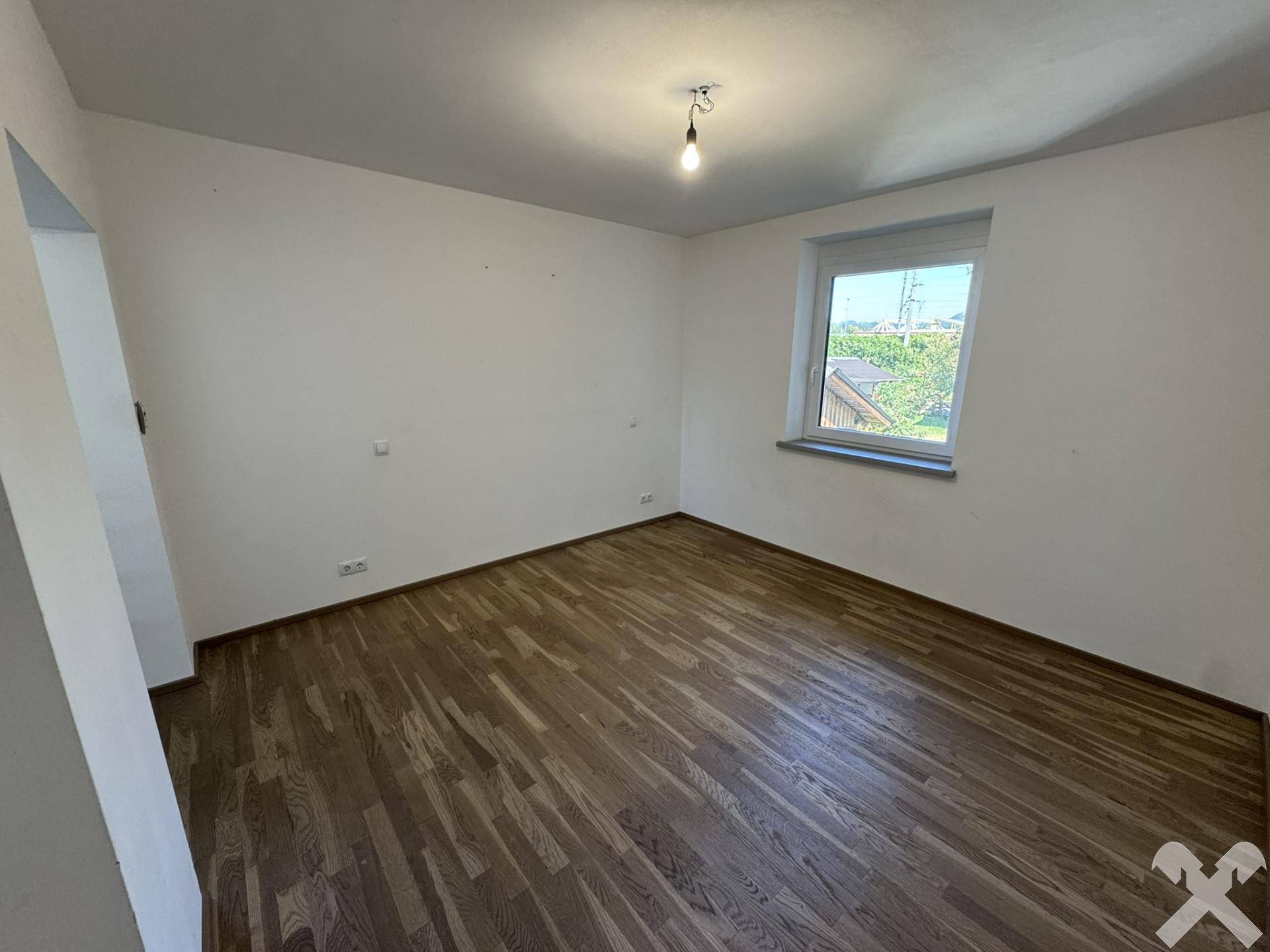 Helles, leeres Zimmer mit Holzboden und einem Fenster mit Blick ins Grüne.