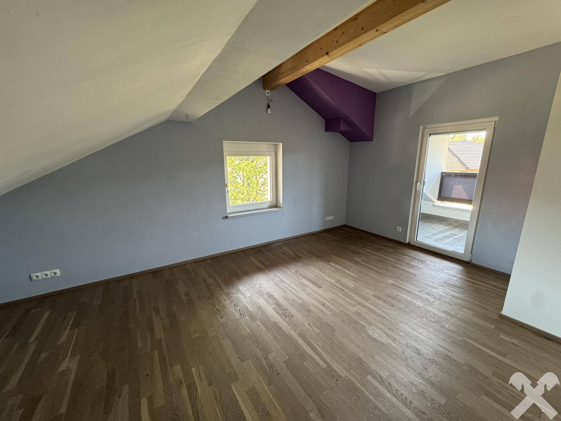 Geräumiges Dachzimmer mit Holzboden, sichtbarem Holzbalken und Zugang zum Balkon.