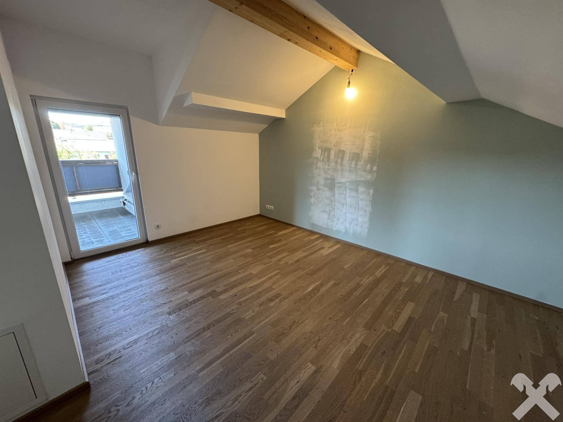 Helles Dachzimmer mit Holzboden und Zugang zu einem Balkon, teilweise farbig gestrichen.