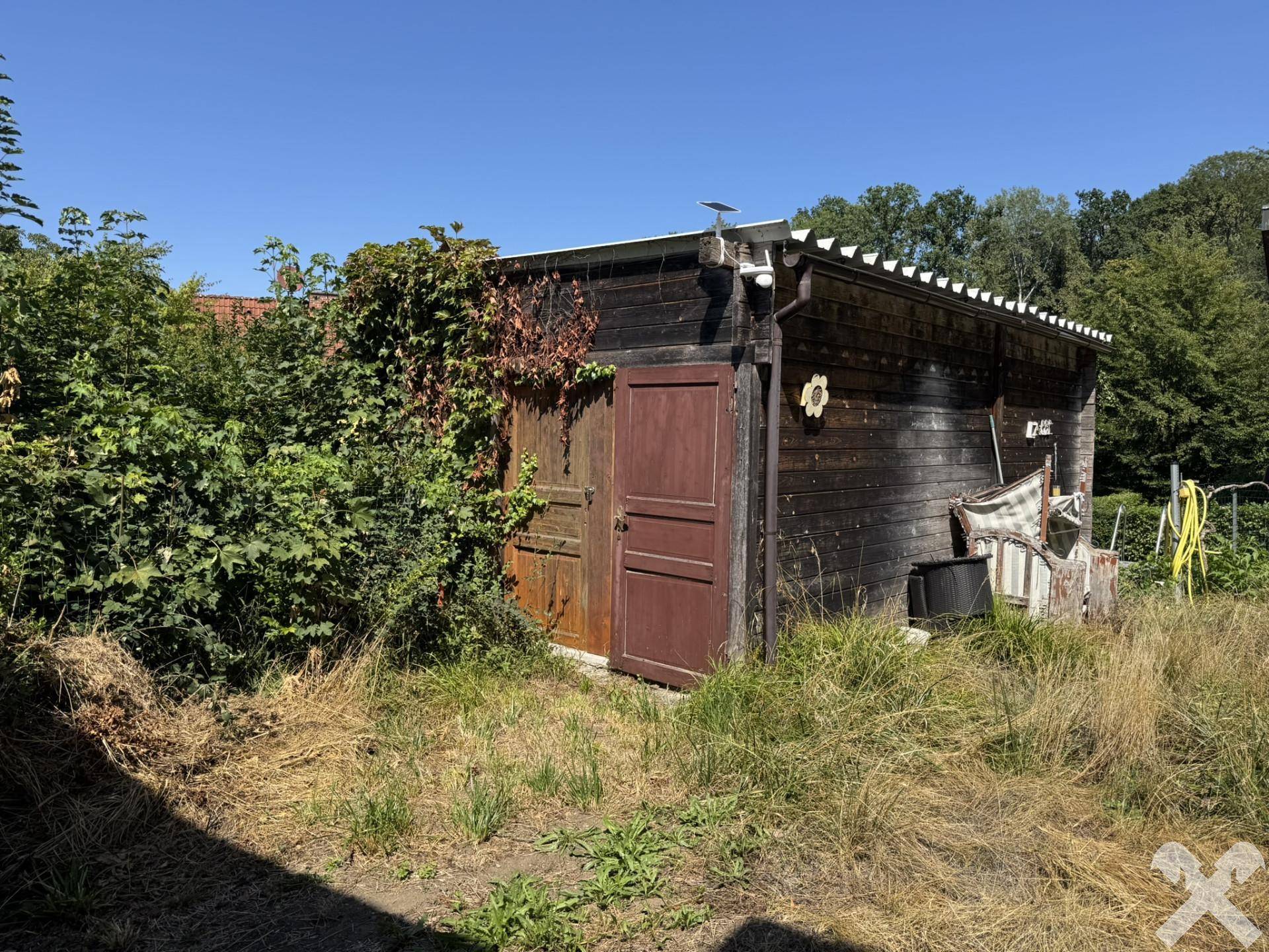 Rustikaler Holzschuppen im Garten, umgeben von üppiger Vegetation und hohem Gras.