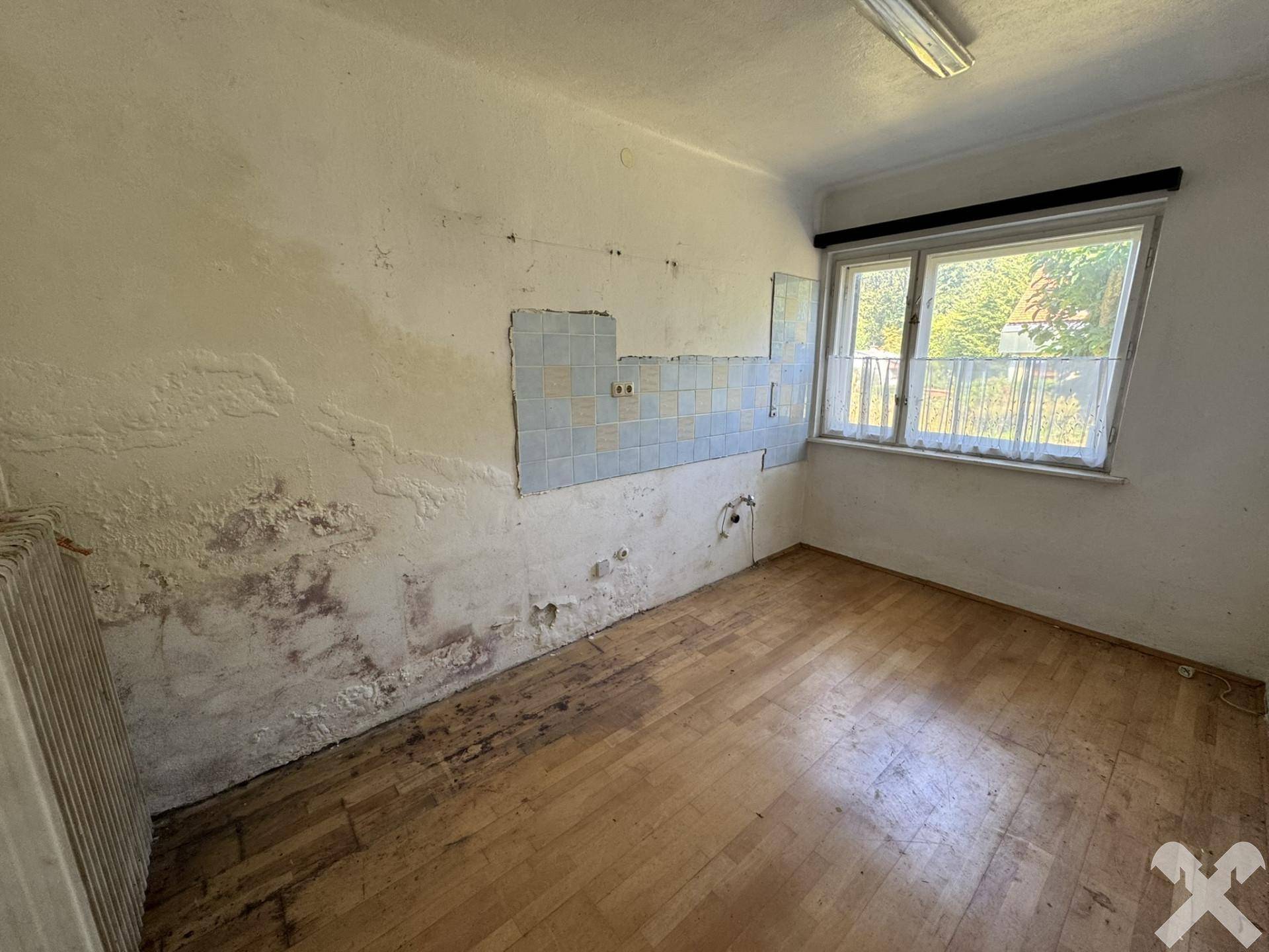 Alte Küche mit beschädigten Wänden, Fliesenresten und einem Fenster, das renovierungsbedürftig ist.