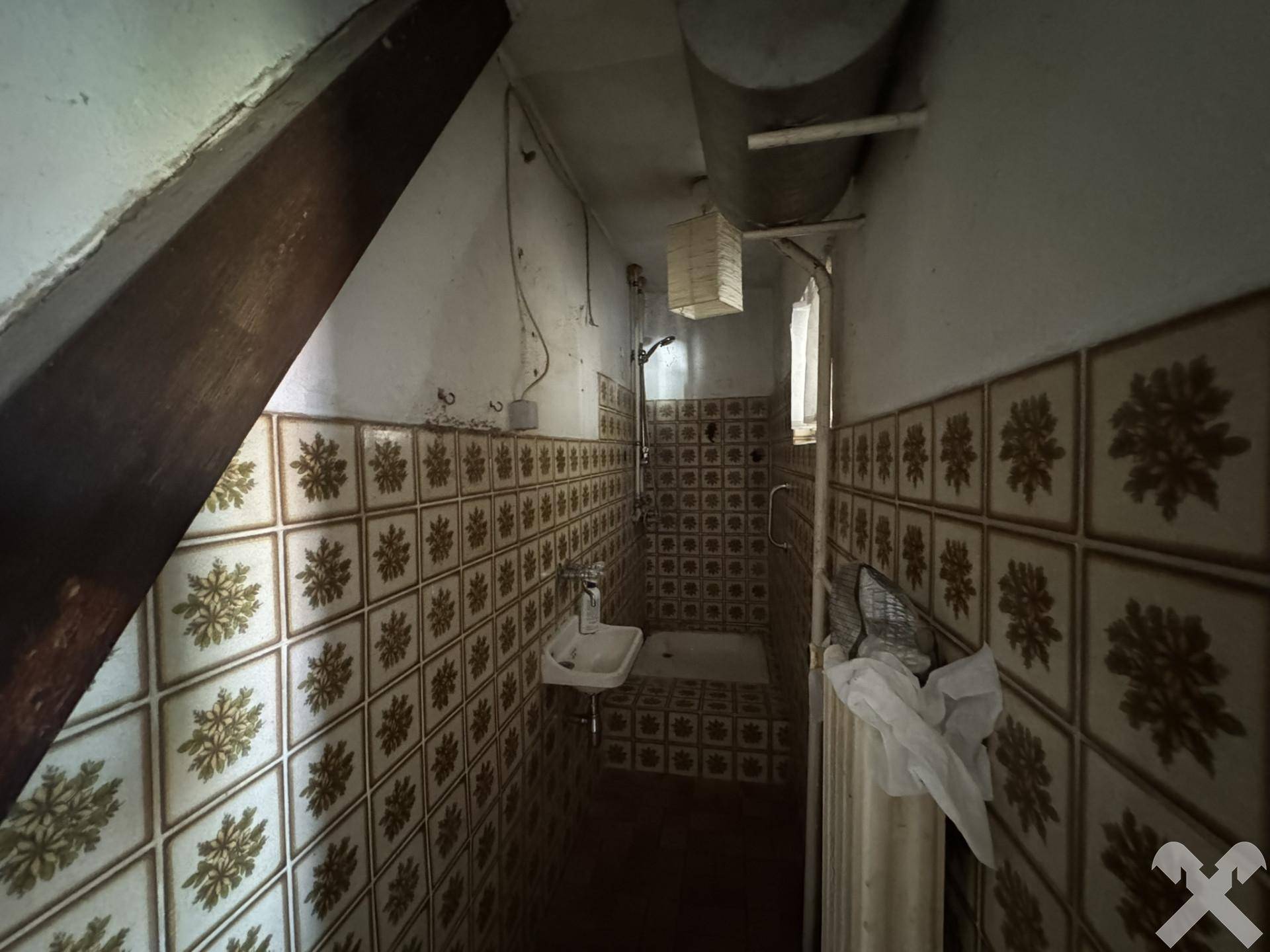 Enges Badezimmer mit Dusche und gemusterten Fliesen unter einer Dachschräge, renovierungsbedürftig.