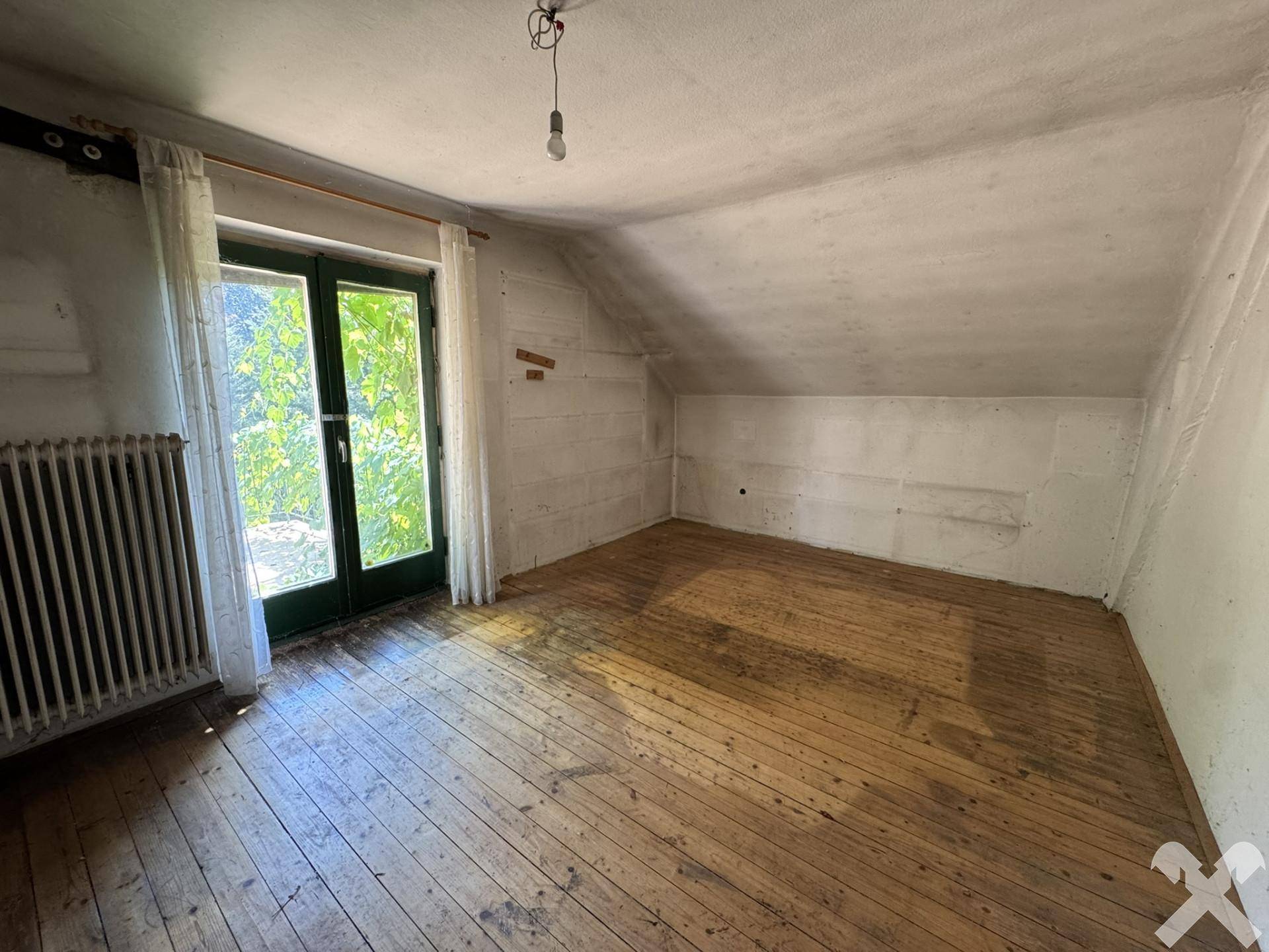 Helles Dachzimmer mit Holzboden, Heizkörper und Zugang zu einem Außenbereich, mit viel Tageslicht.