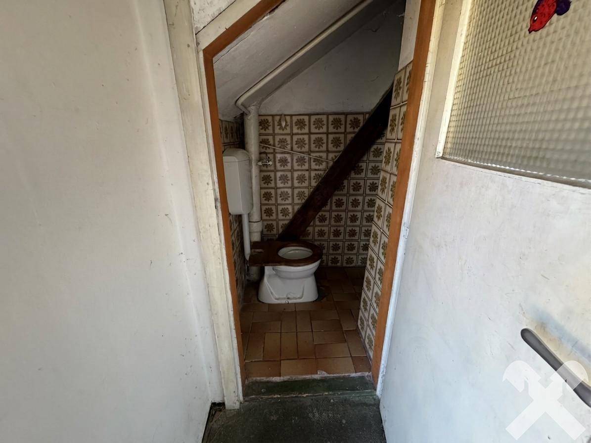 Kleines WC mit Toilette und gemusterten Fliesen unter einer Dachschräge, renovierungsbedürftig.