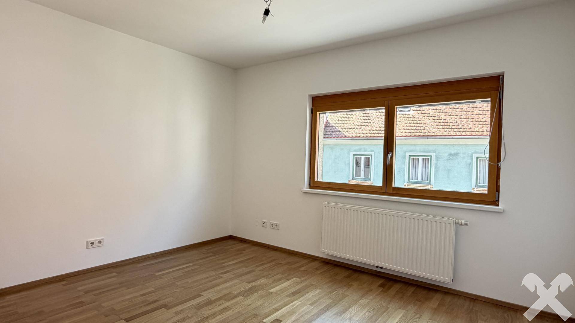 Leeres Schlafzimmer mit Holzparkettboden und einem großen Fenster, das den Raum erhellt.