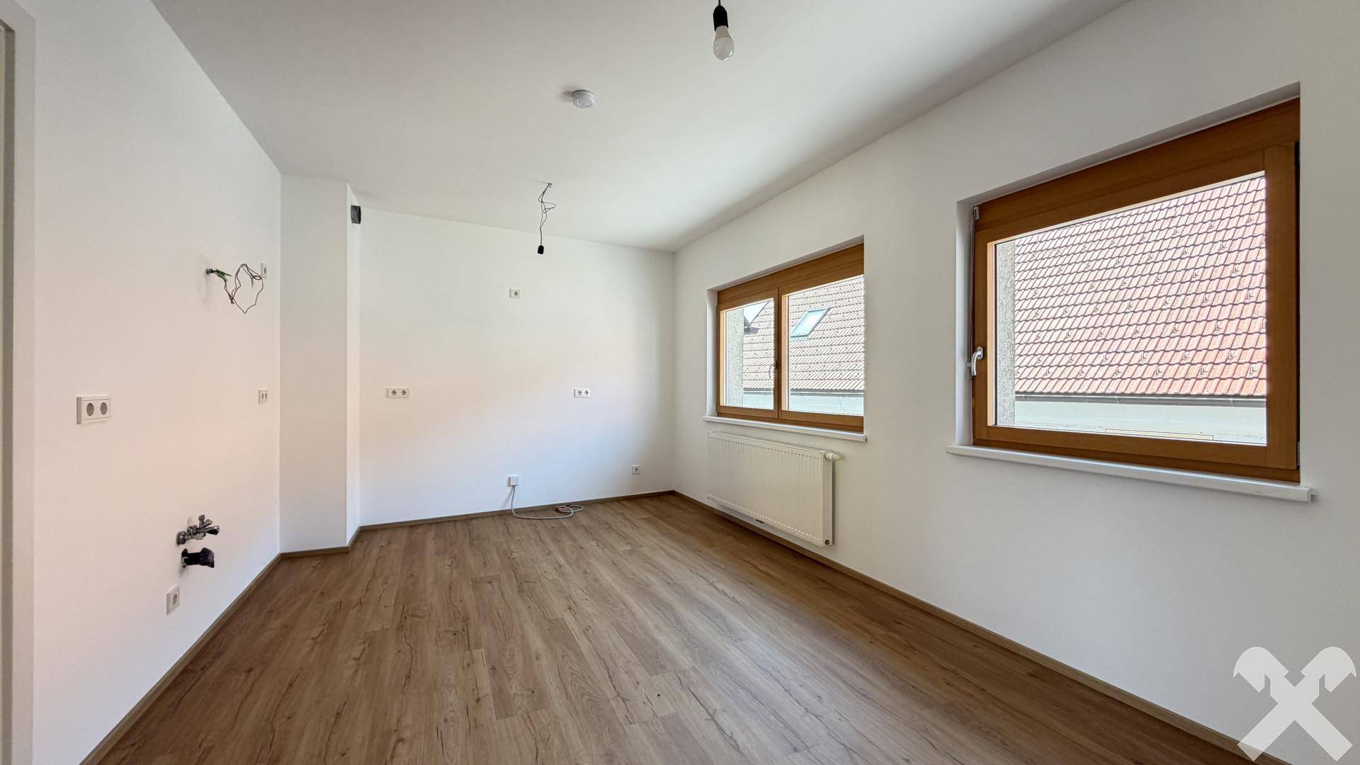 Leerer Küchenraum mit Anschlüssen, Laminatboden und Fenstern, bereit für die Installation.