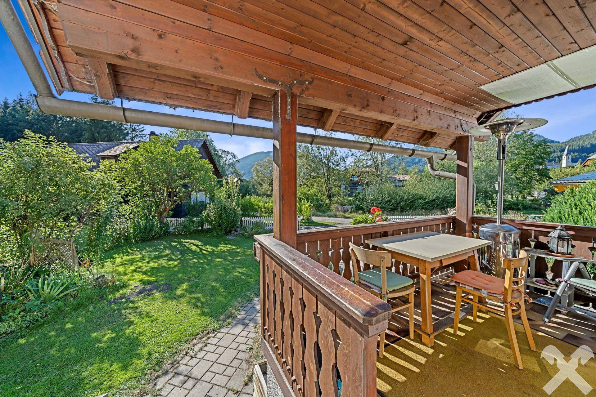 Überdachter Balkon mit Holzbalken, Sitzgelegenheiten und Blick auf den grünen Garten.