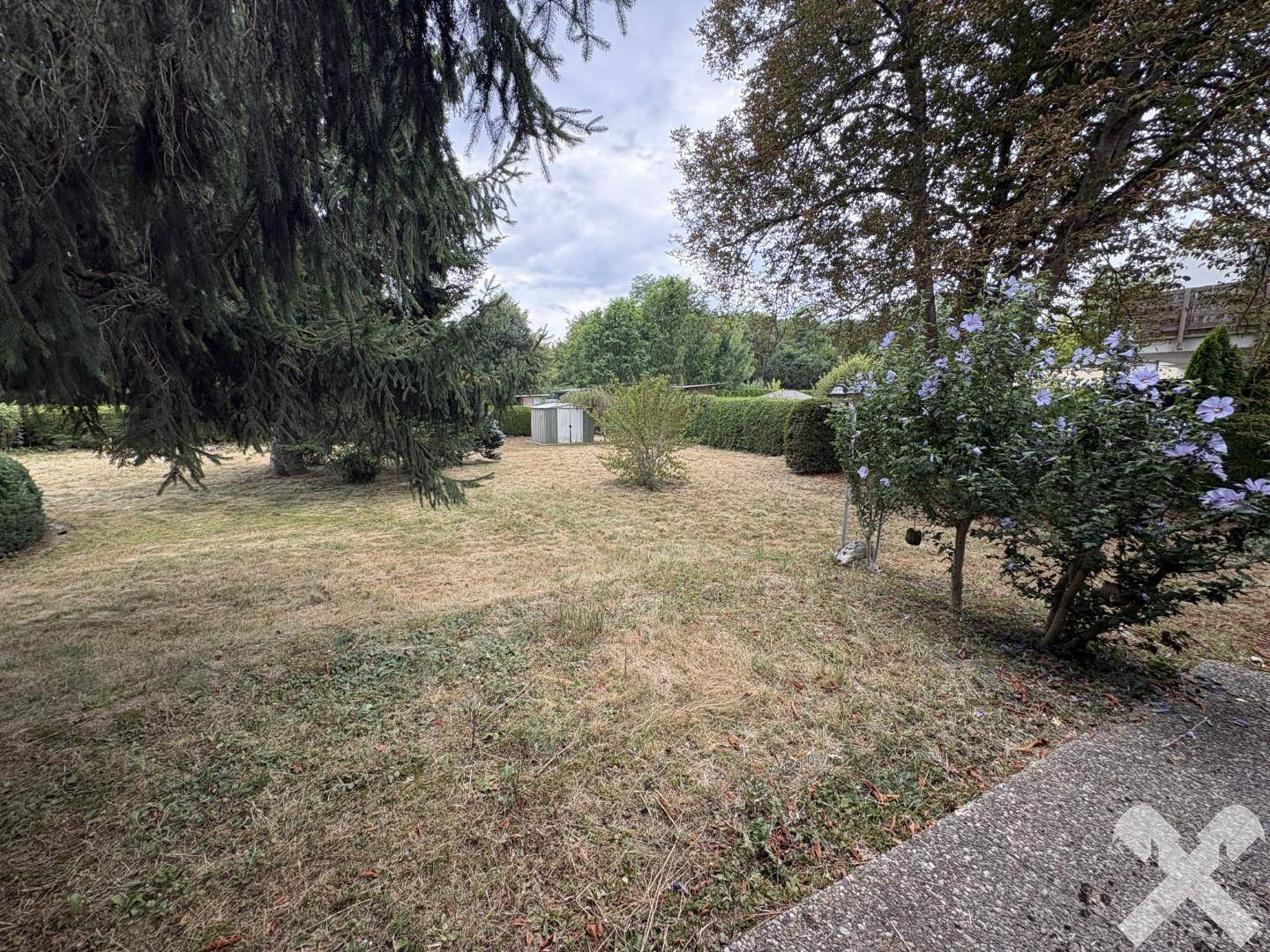 Weitläufiger Gartenbereich mit Rasenfläche, Sträuchern und Bäumen unter bewölktem Himmel.