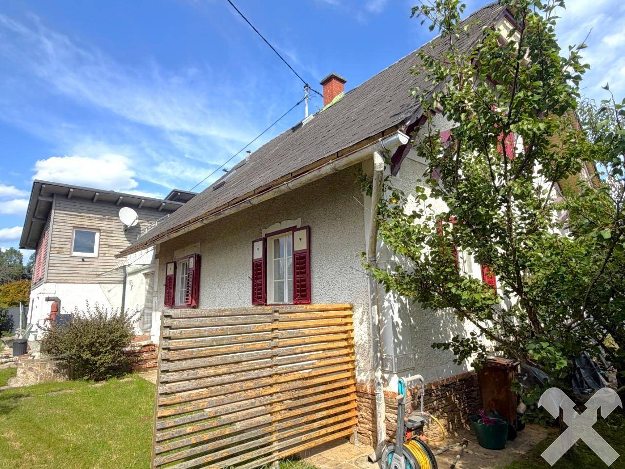 Seitenansicht des älteren Hausteils mit roten Fensterläden und einem Holzzaun im Vordergrund.