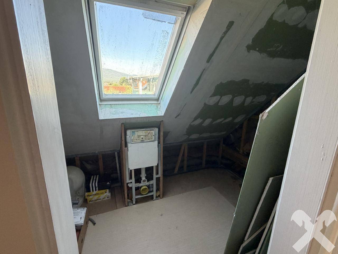 Dachbodenbereich im Rohbau mit sichtbaren Installationen und einem Dachfenster.