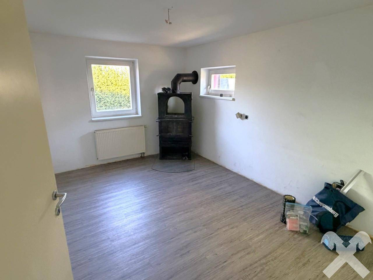 Einfacher Raum mit Holzofen, Laminatboden und zwei Fenstern.