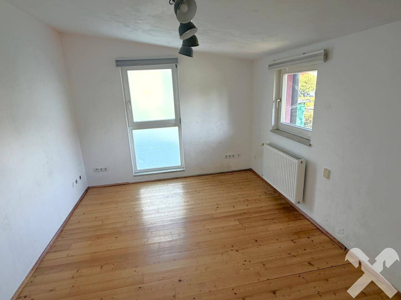 Helles Zimmer mit Holzparkettboden, weißen Wänden und zwei Fenstern.