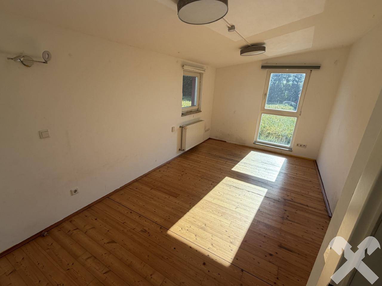 Einfaches Zimmer mit hellen Wänden, Holzdielenboden und viel Tageslicht durch zwei Fenster.