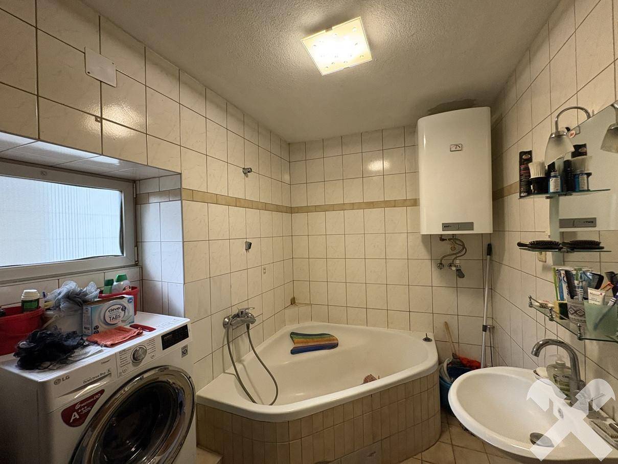 Badezimmer mit Eckbadewanne, Waschmaschine, Waschbecken und hell gefliesten Wänden.