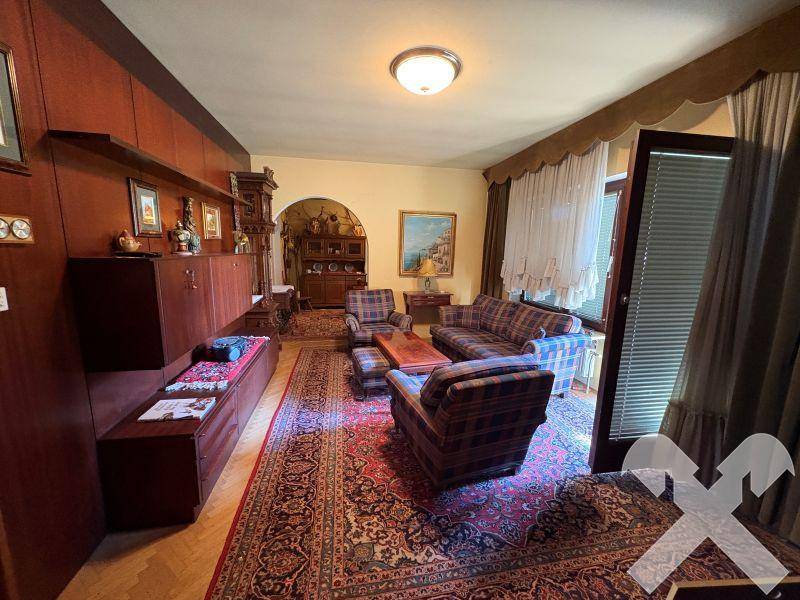 Geräumiges Wohnzimmer mit dunklen Holzmöbeln, Teppichboden und Zugang zum Außenbereich.