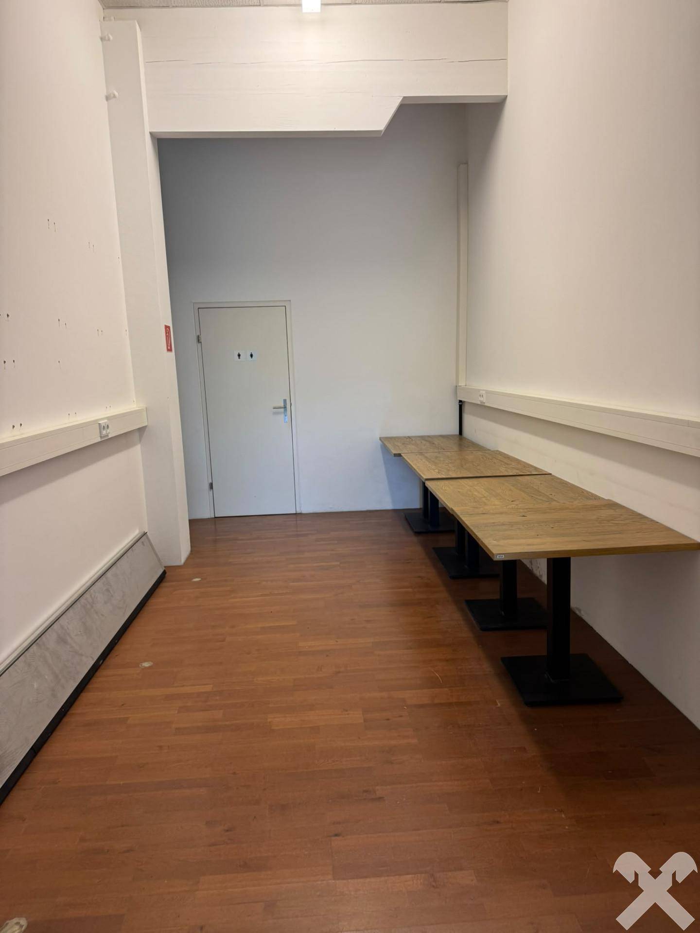 Schmaler Flur oder Wartebereich mit Holzdielenboden, weißen Wänden und an der Wand befestigten Holztischen.