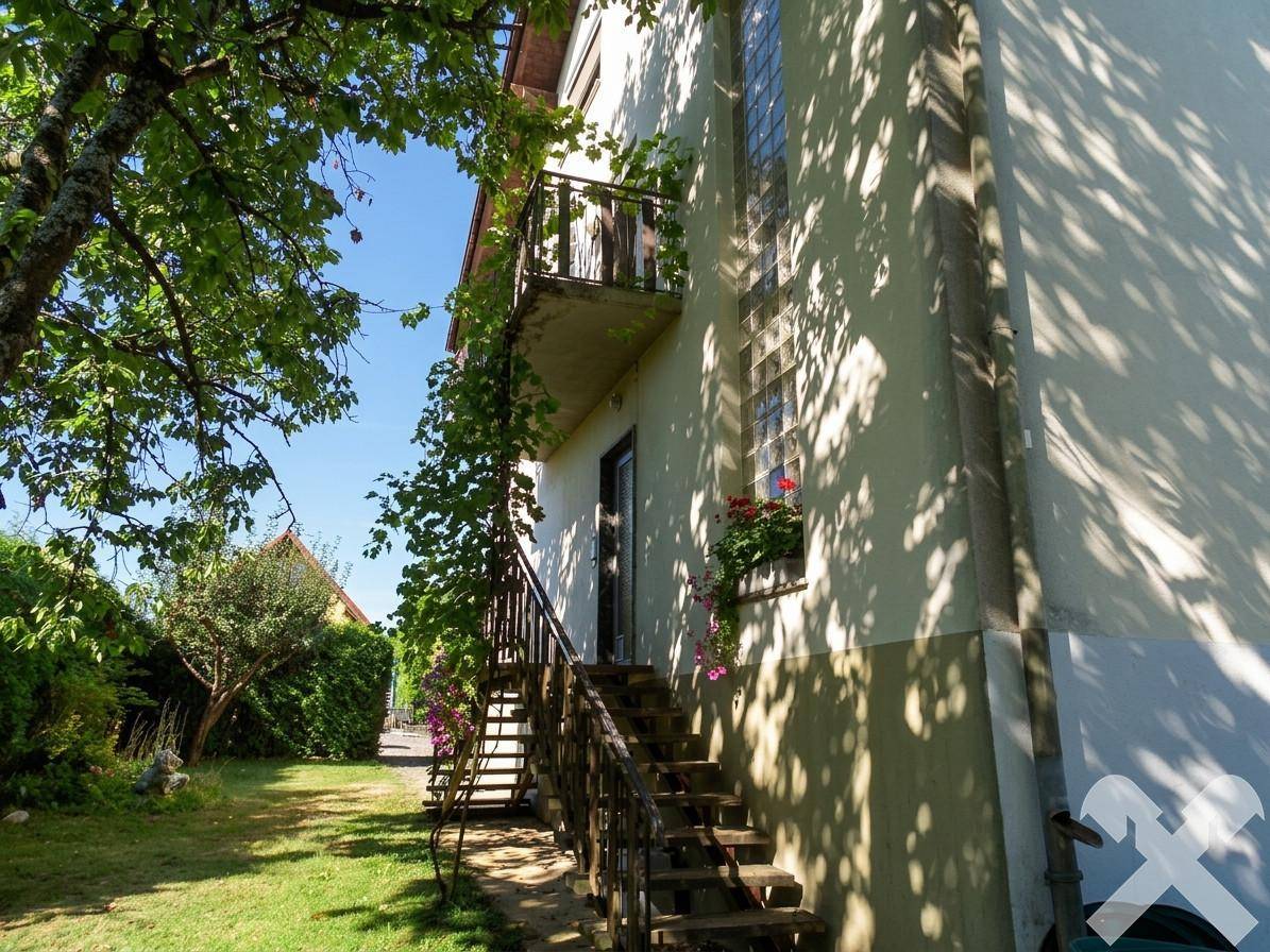 Seitliche Ansicht des Hauses mit Außentreppe, üppigem Grün und sonnigem Gartenbereich.