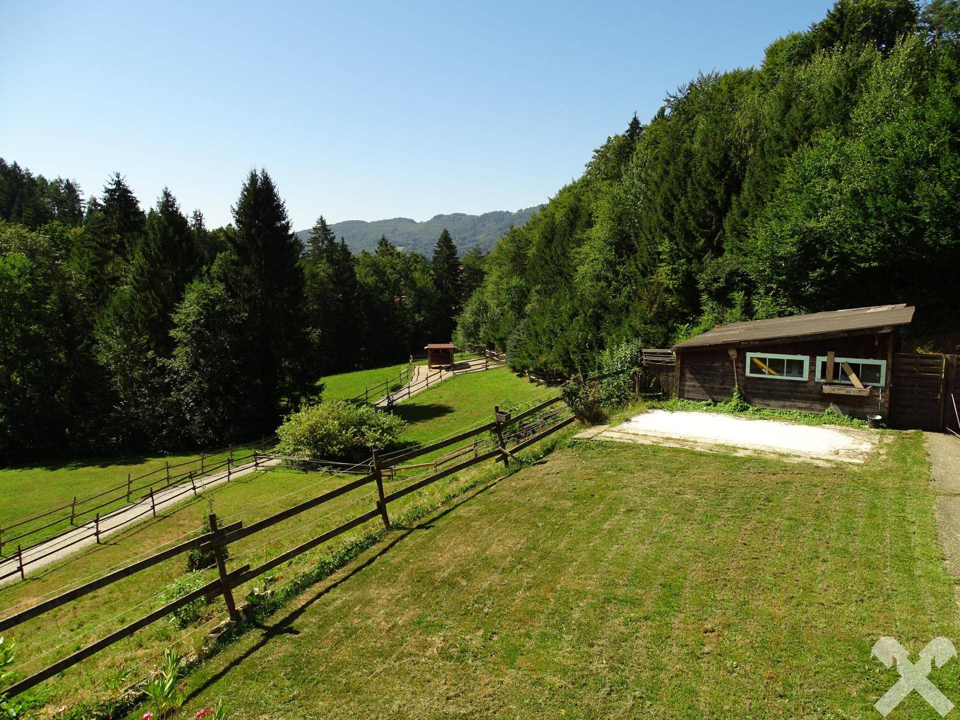 Weitläufige Grünflächen mit Holzzäunen und einem kleinen Holzschuppen vor bewaldeter Hügellandschaft.