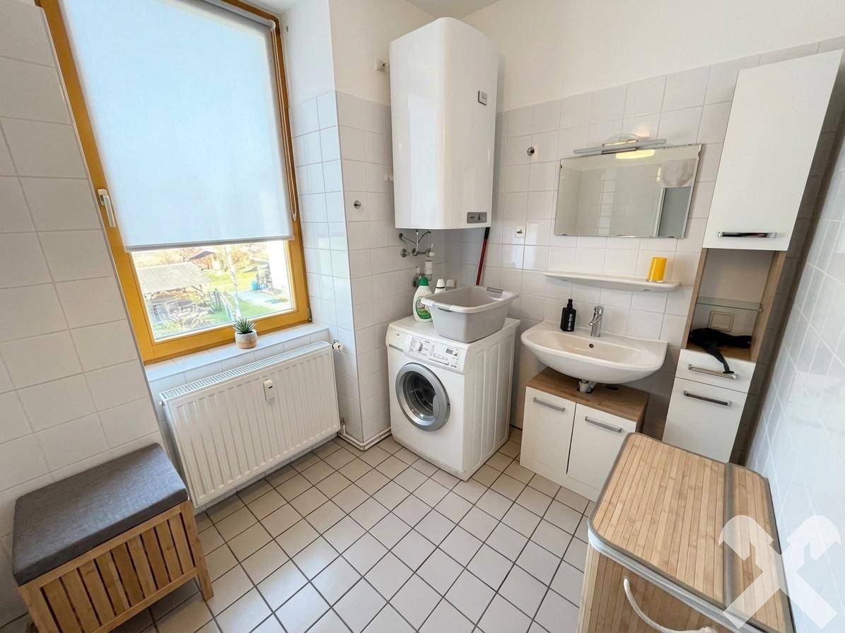 Helles Badezimmer mit Fliesenboden, Waschmaschine, Waschbecken und Fenster.