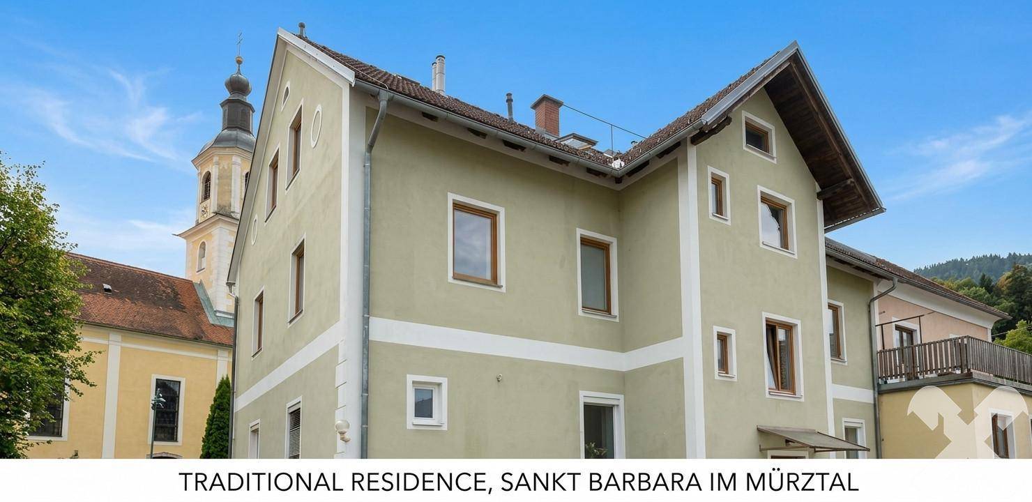 Historisches Wohngebäude mit heller Fassade und Blick auf eine Kirche sowie grüne Hügel.