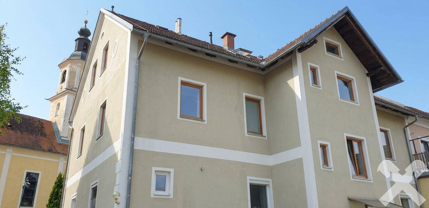 Seitenansicht des Gebäudes mit heller Fassade und einem Kirchturm im Hintergrund unter blauem Himmel.