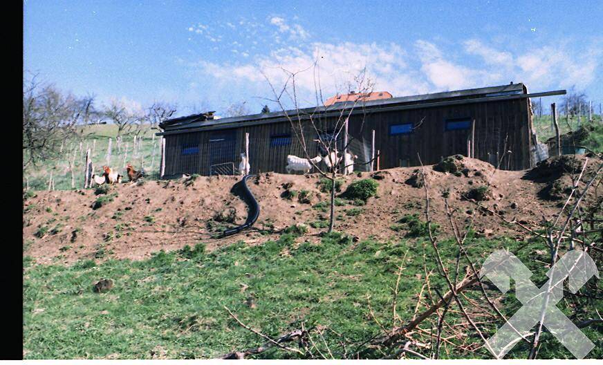 Ländliche Szene mit einem Holzschuppen auf einem Hügel, umgeben von grünen Wiesen und Bäumen.