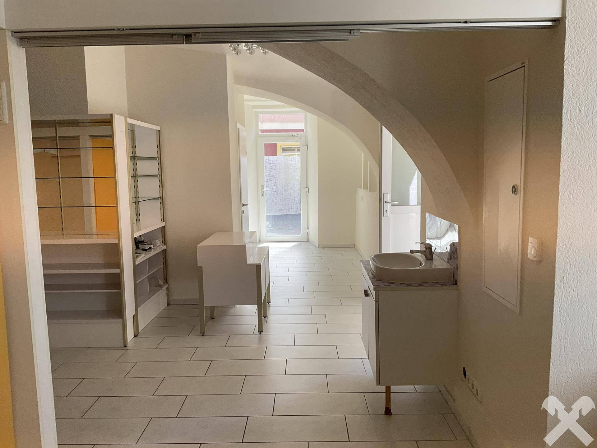 Blick durch einen Bogen in einen weiteren Raum mit Waschbecken, Vitrine und Fliesenboden, der offen gestaltet ist.