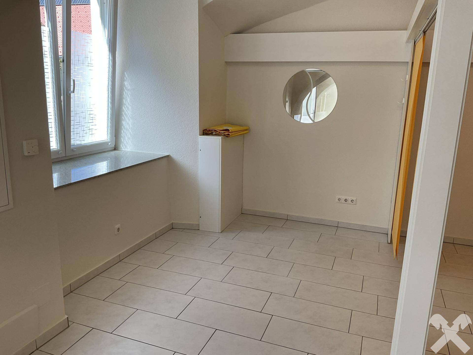Heller Raum mit Fliesenboden, einem Fenster mit Fensterbank und einem runden Wandspiegel, der das Licht reflektiert.