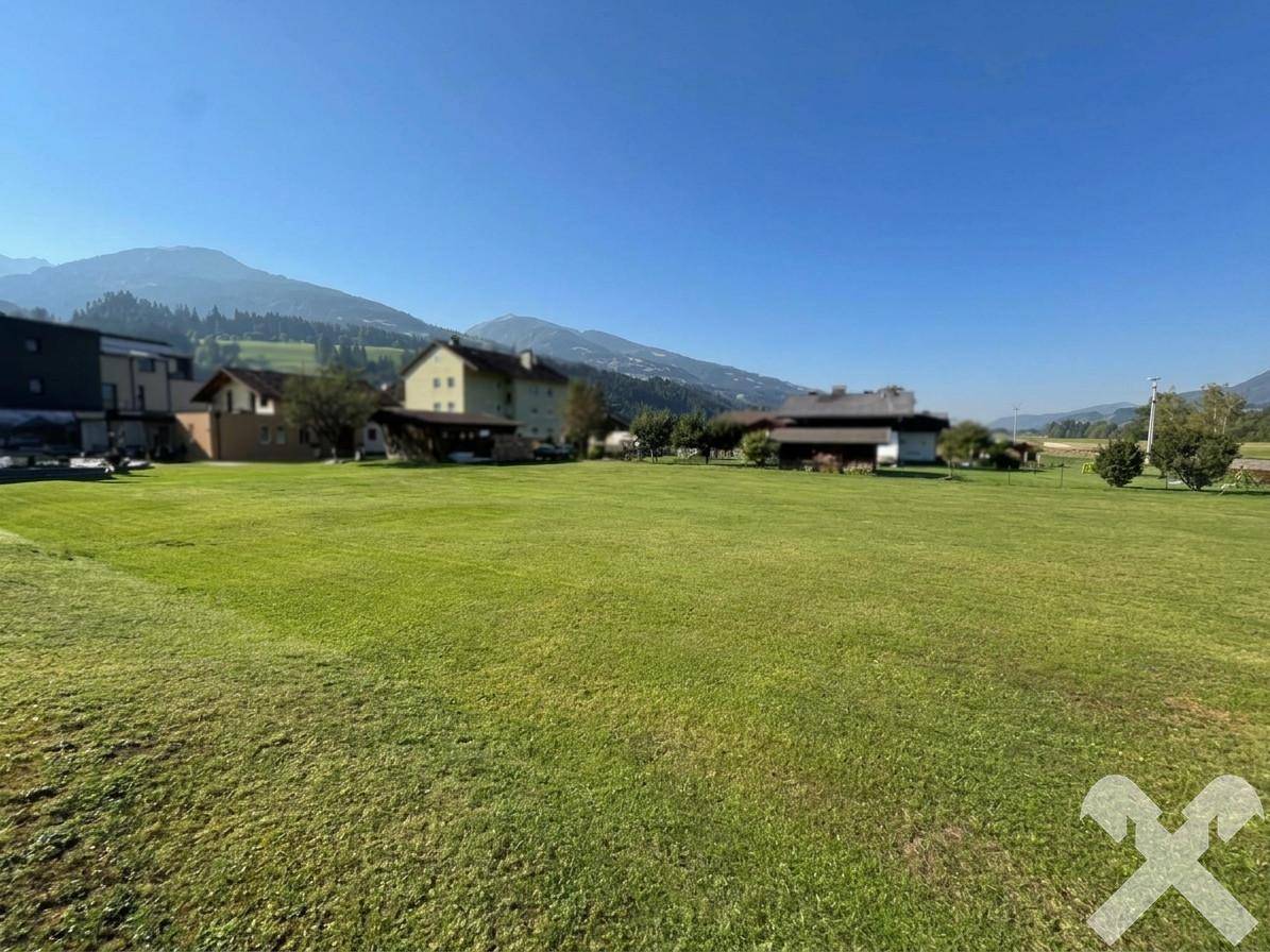 Weitläufige grüne Fläche mit mehreren Häusern und der majestätischen Bergkette im Hintergrund bei Sonnenschein.