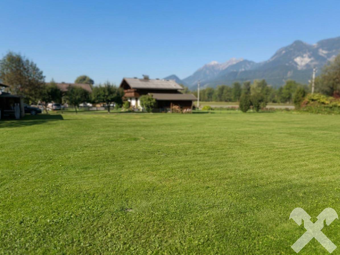 Idyllische Wiesenlandschaft mit traditionellem Haus und Bergblick unter blauem Himmel an einem hellen Tag.
