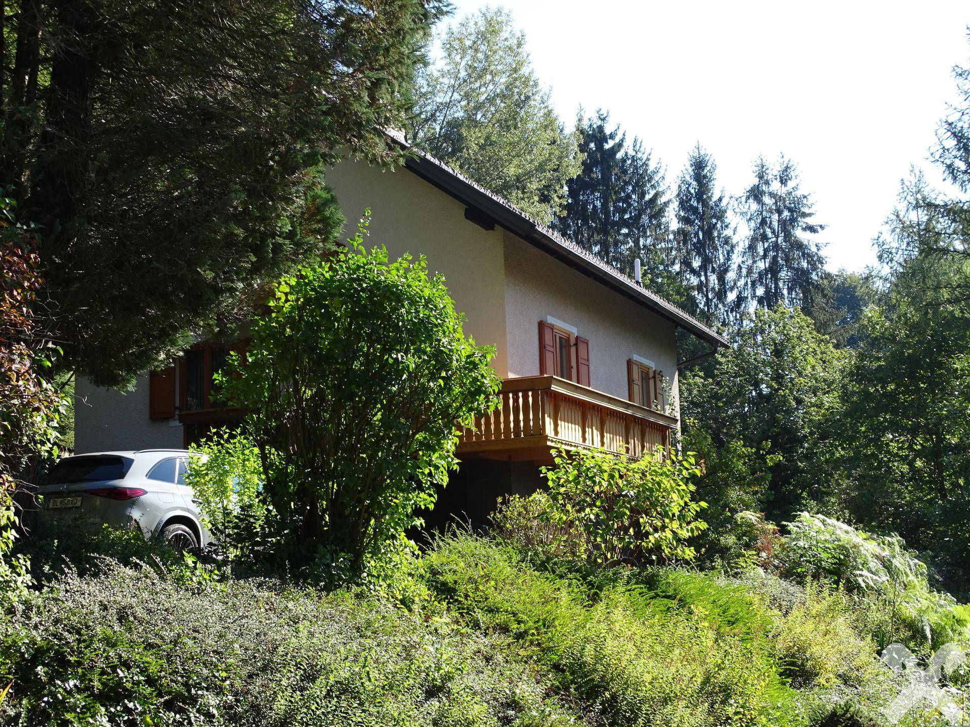 Seitenansicht des Hauses mit einem hölzernen Balkon, umgeben von dichtem Grün und Bäumen.