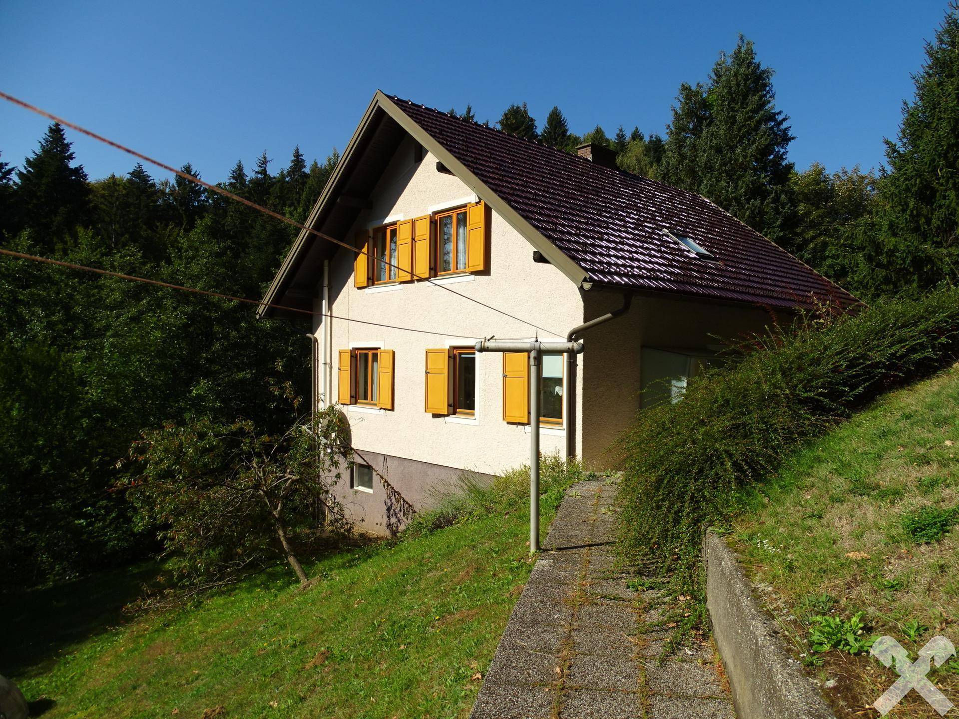 Haus auf einem Hanggrundstück mit gelben Fensterläden und rotem Ziegeldach, umgeben von Natur.