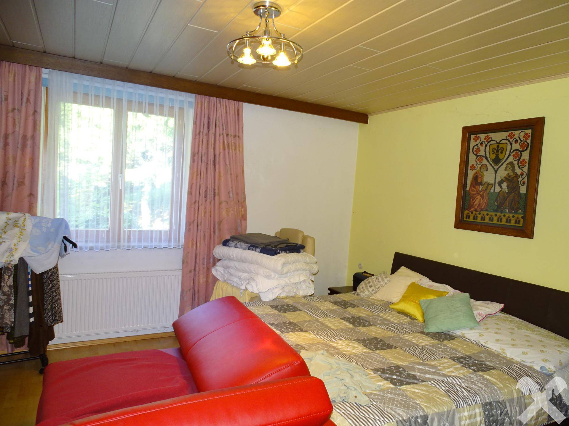 Schlafzimmer mit Doppelbett, rotem Sofa und Fenster, das viel Tageslicht hereinlässt.