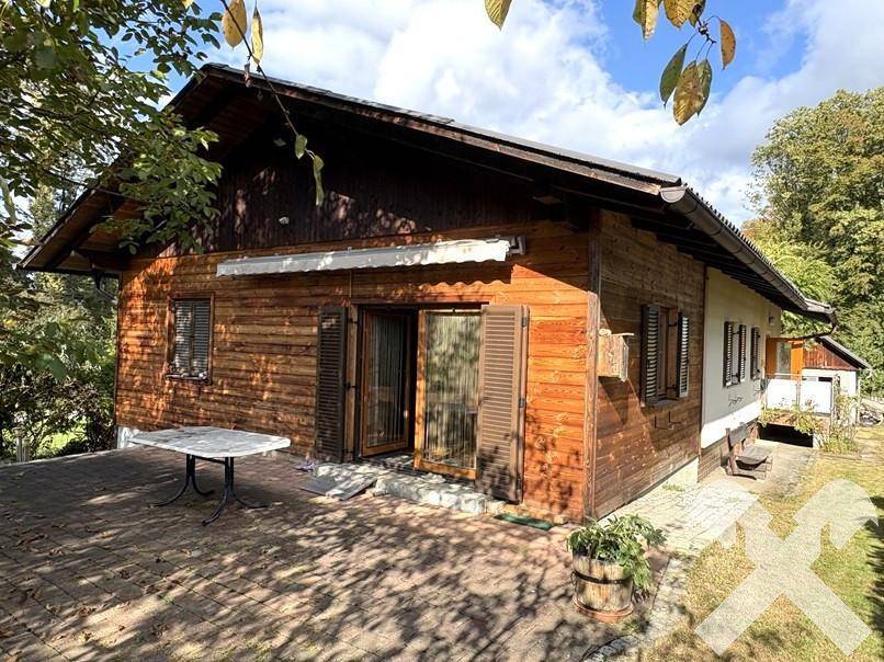 Rustikales Holzhaus mit Terrasse und Blick ins Grüne, umgeben von Bäumen.