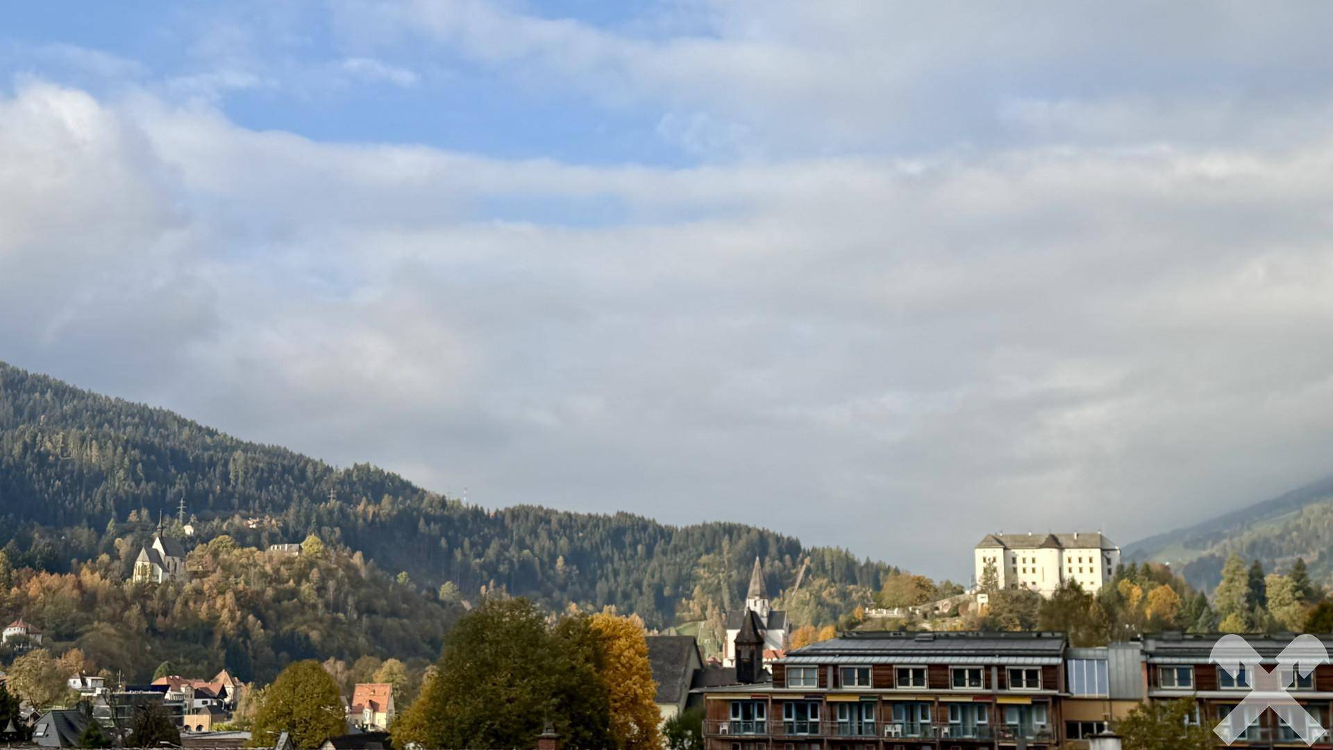Panoramablick auf eine bergige Landschaft mit Häusern und bewaldeten Hügeln unter bewölktem Himmel.