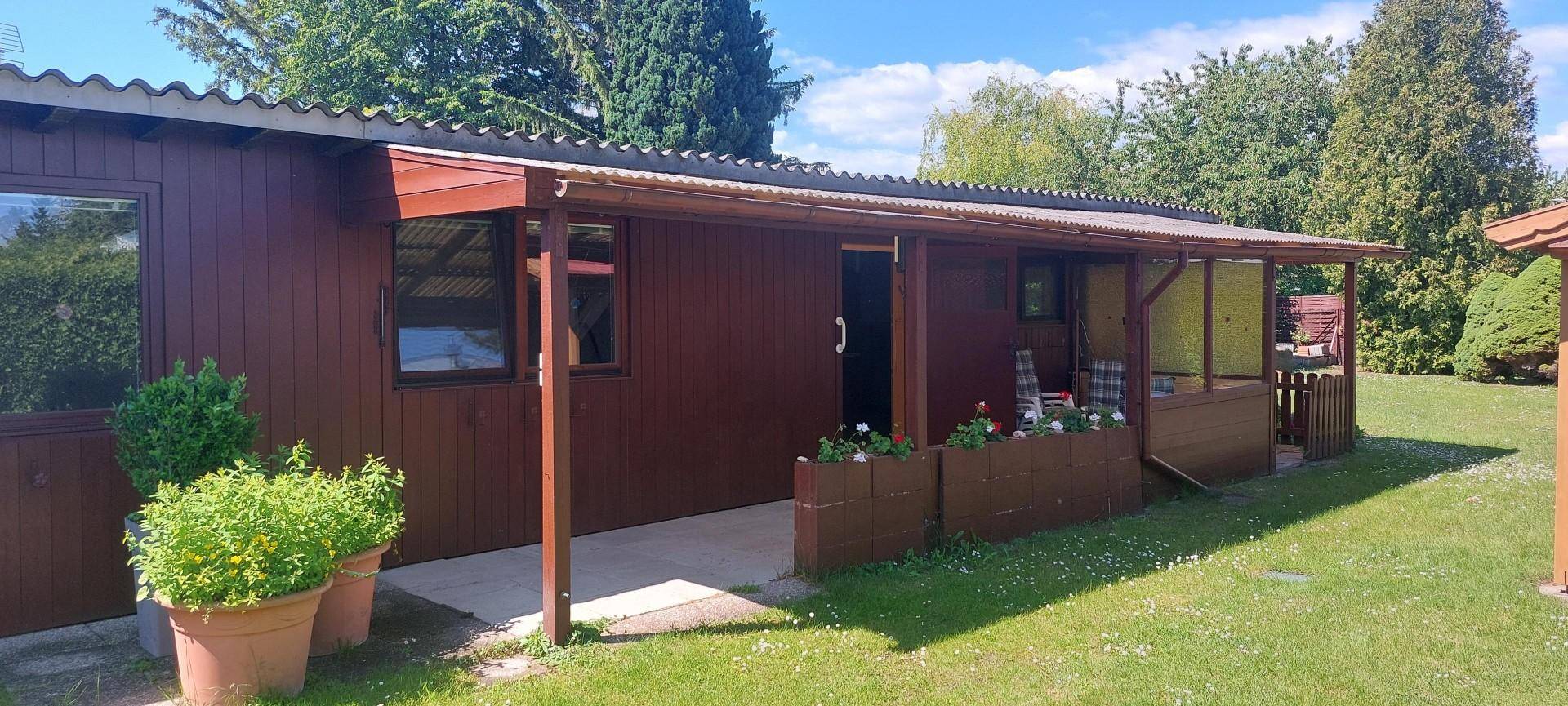 Braunes Holzhaus mit überdachter Terrasse und Pflanzen im Vorgarten unter blauem Himmel.