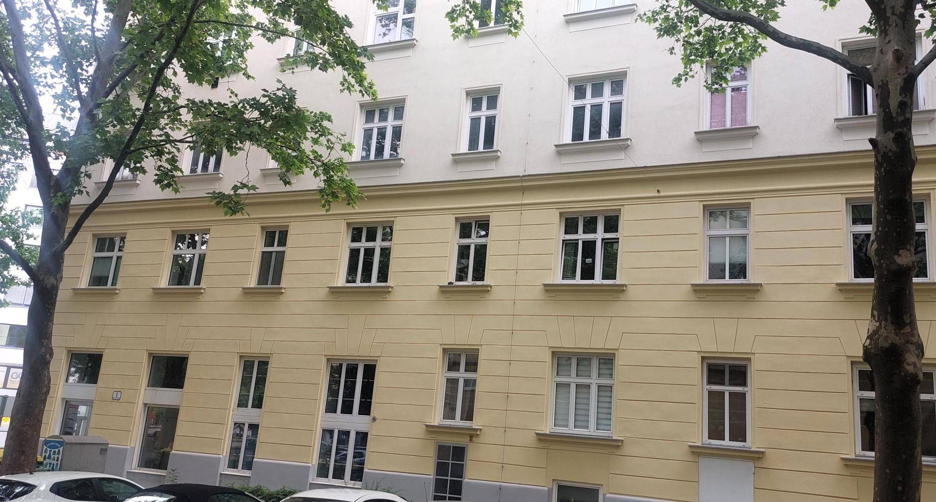 Seitenansicht eines mehrstöckigen Gebäudes mit vielen Fenstern und Bäumen im Vordergrund.